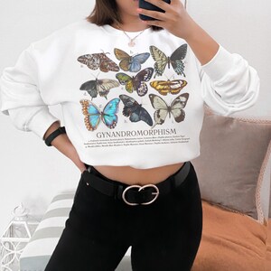 May include: White sweatshirt featuring a collection of butterfly illustrations with the word "GYNANDROMORPHISM" printed below. The butterflies are in various colors and patterns. The sweatshirt is paired with black pants and a black belt with a gold buckle.