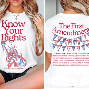 Puede incluir: Camiseta blanca con el texto rojo "Know Your Rights", con una ilustración roja, blanca y azul de la Estatua de la Libertad. La parte trasera de la camiseta muestra "The First Amendment" con una pancarta de banderas.