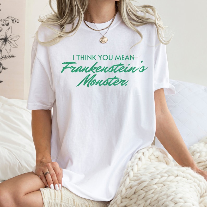 May include: White t-shirt with the text "I THINK YOU MEAN Frankenstein's Monster" in green lettering. The shirt is a casual style, suitable for everyday wear. The text is in a handwritten style.