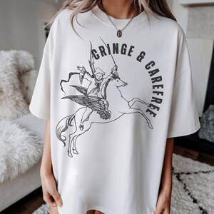 May include: Cream-colored t-shirt with a black graphic of a winged horse ridden by a jester-like figure. The words "CRINGE & CAREFREE" are printed in a semi-circle around the image. The shirt has a relaxed fit.