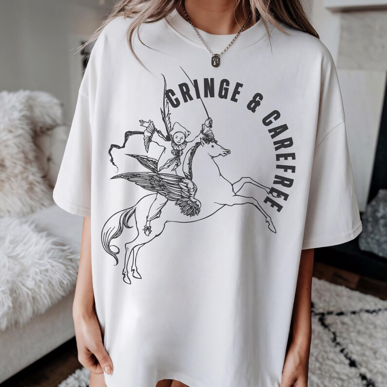 May include: Cream-colored t-shirt with a black graphic of a winged horse ridden by a jester-like figure. The words "CRINGE & CAREFREE" are printed in a semi-circle around the image. The shirt has a relaxed fit.