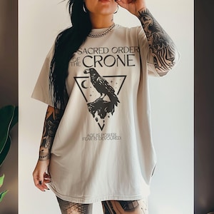 May include: A light beige t-shirt with a graphic design featuring a raven perched on a rock within a triangle. The text reads "SACRED ORDER OF THE CRONE" and "AGE IS POWER FEAR IS DEVOURED".