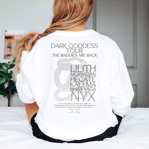 May include: White long-sleeve sweatshirt with a black graphic print of a snake and text that reads "Dark Goddess Tour The Baddies Are Back" and lists names like Lilith, Morrigan, Hecate, Kali Ma, Baba Yaga, and Nyx. The text also includes "It's the worldwide tour spend an untamed and transformative evening with the greatest dark feminine hits like independence, assertion, intuition and self worth. DOC"