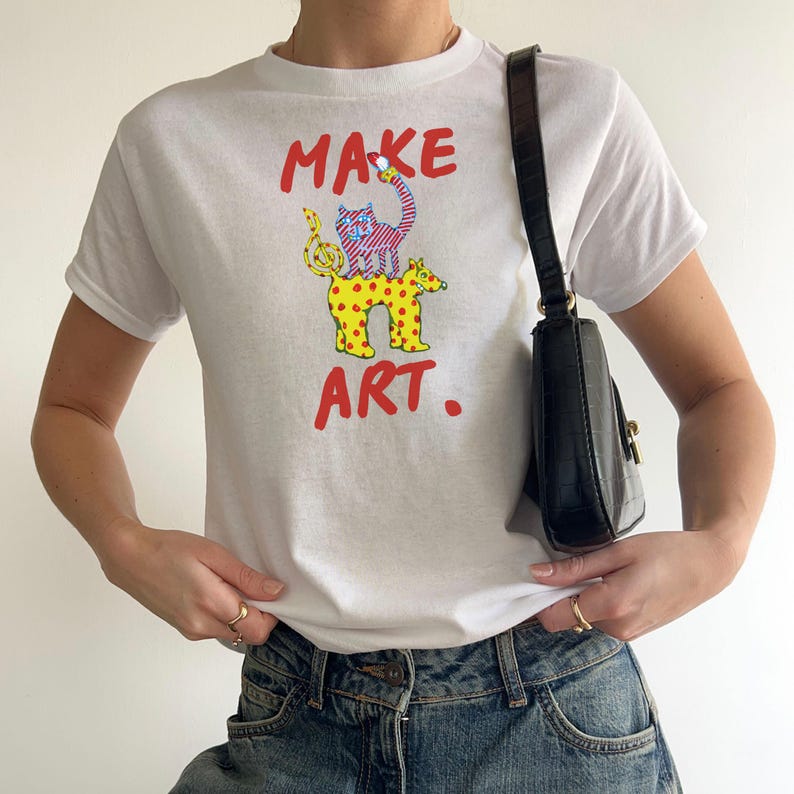 May include: White t-shirt with the words "MAKE ART." in red, featuring a whimsical illustration of a cat and a spotted animal. A black handbag hangs from the shoulder. The model is wearing blue jeans.