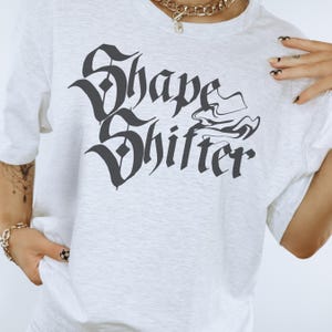 Pode incluir: Camiseta branca com texto preto que diz "Shape Shifter" em uma fonte gótica.