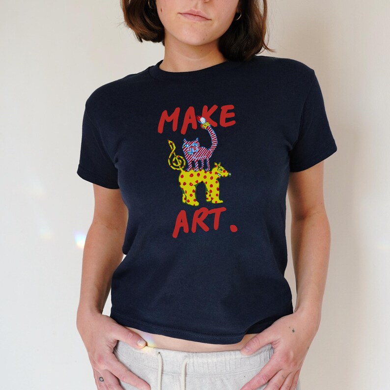 May include: Navy blue t-shirt with the words "MAKE ART." in red. The shirt features a whimsical illustration of a cat and a spotted animal. The cat is pink and the spotted animal is yellow.