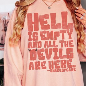 May include: A light pink sweatshirt with the text "HELL IS EMPTY AND ALL THE DEVILS ARE HERE." in a bold, red font. The quote is attributed to Shakespeare. The sweatshirt has a crew neck and long sleeves.