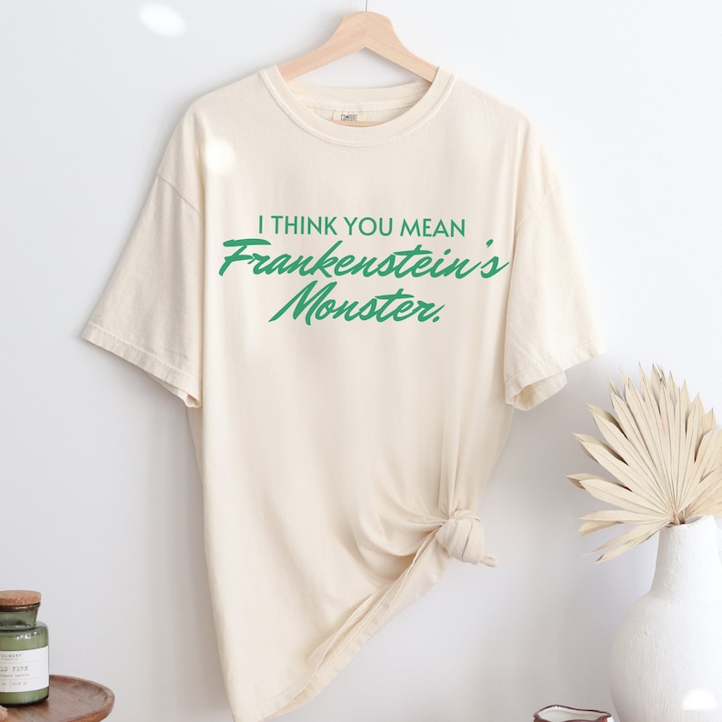 May include: Cream-colored t-shirt with the text "I THINK YOU MEAN Frankenstein's Monster" in green. The shirt is hanging on a wooden hanger. The shirt is knotted at the bottom.