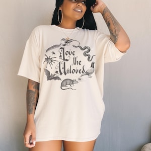 May include: Off-white t-shirt with a graphic of a spider, snake, and bat, encircling the words "Love the Unloved". The tee has short sleeves and a loose fit. The design is in black.