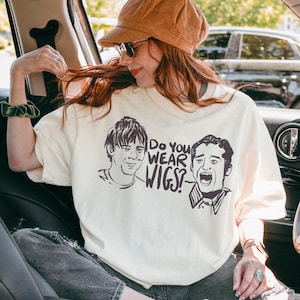 May include: Cream-colored t-shirt featuring a graphic of two men with the text "Do You Wear Wigs?" in black. The shirt is worn by a person in a car, wearing a brown hat and sunglasses.