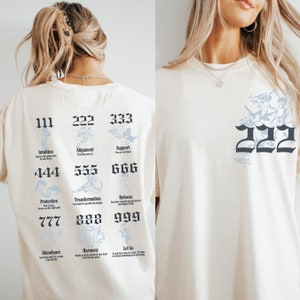 以下が含まれることがあります： 天使の羽根と数字のグラフィックプリントが施された白いTシャツ。111、222、333、444、555、666、777、888、999の数字がシャツの背中にプリントされており、それぞれに対応する単語またはフレーズが添えられています。単語は、直感、アライメント、サポート、プロテクション、トランスフォーメーション、ベロカス、アバンダンス、ハーモニー、レットゴーです。