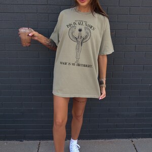 May include: An olive green oversized t-shirt with the text "BETWEEN ALL NAMES ISIS IN ALL NAMES MAGIC IS MY BIRTHRIGHT" and a graphic of a figure with outstretched wings. The shirt is worn with white sneakers.