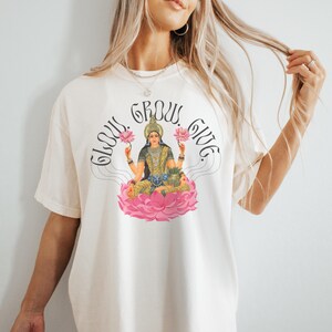 May include: A cream-coloured t-shirt with a graphic of a seated deity on a pink lotus flower, holding pink lotus flowers. The words "Glow, Grow, Give" are arched above the image in black script. The shirt has short sleeves.