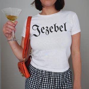 May include: A white t-shirt with the word "Jezebel" printed in black gothic lettering. The shirt is being worn by a person with a black and white checkered skirt and an orange crossbody bag.