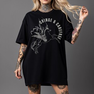 May include: Black oversized t-shirt with a white graphic of a winged horse ridden by a figure with a sword. The text "CRINGE & CAREFREE" is arched around the image. The shirt has short sleeves and a relaxed fit.