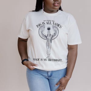 May include: Off-white t-shirt featuring a black graphic of a winged figure with the text "ISIS IN ALL NAMES" and "MAGIC IS MY BIRTHRIGHT." The shirt has short sleeves and a crew neck. The model is wearing blue jeans.