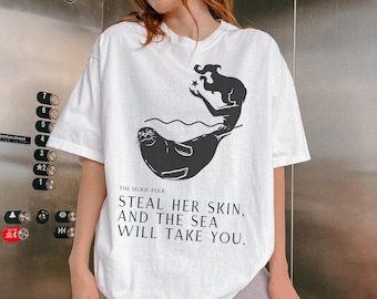 May include: White t-shirt featuring a black graphic of a seal and a mermaid-like figure holding a star. The text "THE SELKIE-FOLK STEAL HER SKIN, AND THE SEA WILL TAKE YOU" is printed below the graphic. The shirt is a casual style.