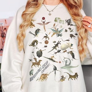 May include: Cream-colored sweatshirt featuring a detailed illustration of fantastical creatures and figures in various poses. The design includes a variety of animals and human-like figures, with the text "Hieronymus Bosch Creatures" printed in a vertical arrangement.