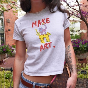 May include: A light gray t-shirt with the words "MAKE ART." in red. The shirt features a whimsical graphic of a cat and dog with musical notes. The cat has a striped pattern, and the dog has yellow spots.