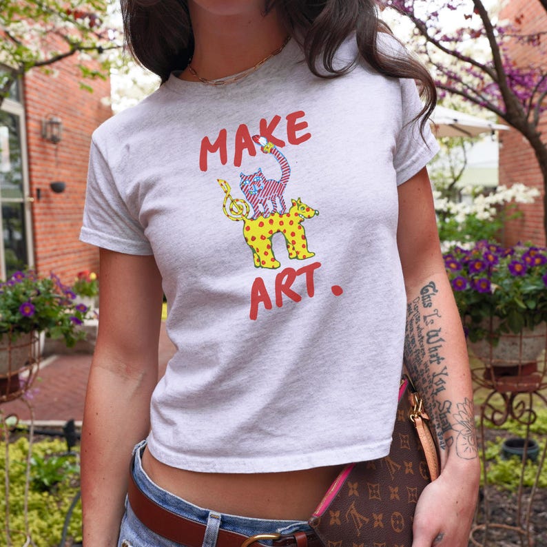 May include: A light gray t-shirt with the words "MAKE ART." in red. The shirt features a whimsical graphic of a cat and dog with musical notes. The cat has a striped pattern, and the dog has yellow spots.