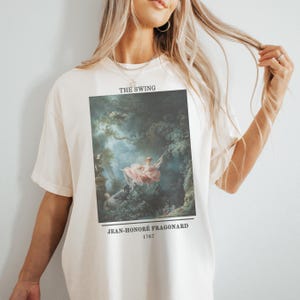 May include: A white t-shirt featuring a print of the painting "The Swing" by Jean-Honoré Fragonard. The painting depicts a woman on a swing in a lush garden setting. The text "THE SWING" is printed above the painting, and the artist's name and the year 1767 are printed below the painting.