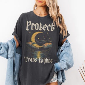 Peut inclure: Un t-shirt gris foncé avec un graphique de croissant de lune et d'étoiles. Le texte "Protect Trans Rights" est imprimé sous le graphique.