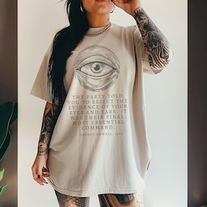 May include: Beige oversized t-shirt with a graphic of an eye inside a circle. The shirt features a quote from George Orwell's "1984" below the graphic. The t-shirt has short sleeves and a loose fit, ideal for everyday wear.