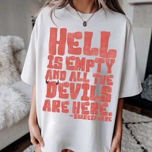 May include: Off-white t-shirt with the text "HELL IS EMPTY AND ALL THE DEVILS ARE HERE." in coral-colored, bubble-style letters. The shirt also features a small devil horn graphic and the name "-SHAKESPEARE" below the quote.