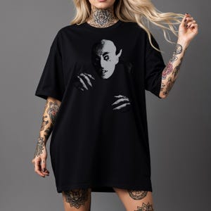 Puede incluir: Camiseta negra con un gráfico en escala de grises de la cara y las manos de un vampiro. El diseño tiene una estética vintage de película de terror. La camiseta es de corte holgado, con mangas cortas.