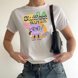 May include: White t-shirt with the text "Sluttiest Slut at the Library" in a colorful design. The graphic includes a cartoon book character with glasses and a pointer. The shirt is paired with blue jeans and a black purse.