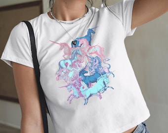 May include: White crop top featuring a cluster of unicorns in shades of blue and pink. The graphic design includes various unicorn poses, creating a whimsical and fantasy-themed image. The shirt has short sleeves and a crew neck.