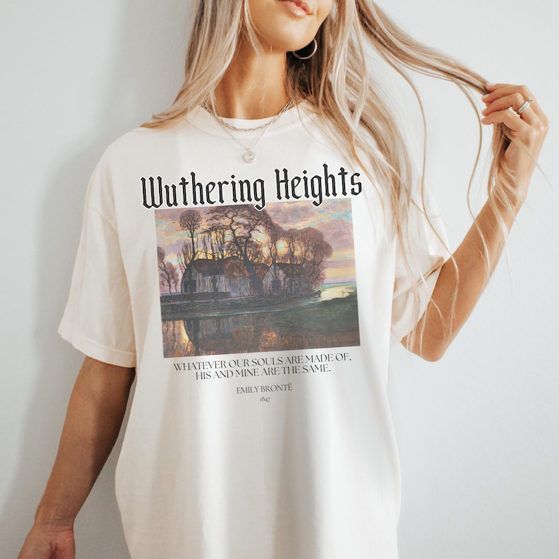 May include: A white t-shirt with a black graphic print of a painting of a house with trees and a lake. The text "Wuthering Heights" is printed above the painting. Below the painting is a quote from the book: "Whatever our souls are made of, his and mine are the same." The quote is attributed to Emily Bronte.