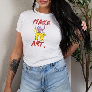 May include: White t-shirt with the words "MAKE ART." in red, featuring a colorful cartoon illustration of two animals. The top animal is pink and blue, and the bottom animal is yellow with brown spots.