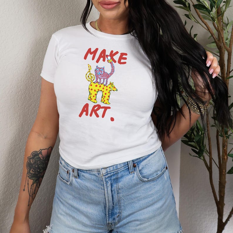 May include: White t-shirt with the words "MAKE ART." in red, featuring a colorful cartoon illustration of two animals. The top animal is pink and blue, and the bottom animal is yellow with brown spots.