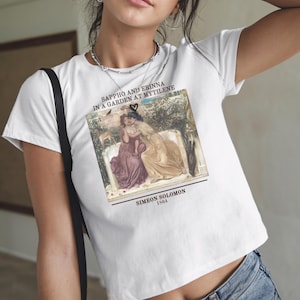 May include: White t-shirt with a framed image of a painting titled "Sappho and Erinna in a Garden at Mytilene" by Simeon Solomon, 1864. The artwork shows two women embracing. A black heart is above the image.