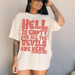 May include: Cream-colored t-shirt with the text "HELL IS EMPTY AND ALL THE DEVILS ARE HERE." in red, with a small devil graphic. The quote is attributed to Shakespeare. The shirt is a casual, oversized fit.
