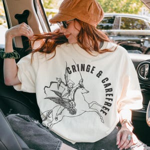 May include: Cream-colored t-shirt with a black graphic of a figure riding a winged horse, and the words "CRINGE & CAREFREE" in a circular design. The shirt is worn by a person in a car, wearing a brown hat and sunglasses.