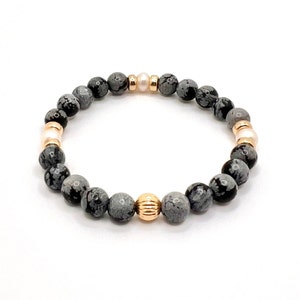 May include: A beaded bracelet featuring gray and black speckled stone beads, interspersed with gold-colored and pale pink beads. The bracelet is strung on an elastic band, showcasing a mix of textures and colors. The bracelet is designed for everyday wear.