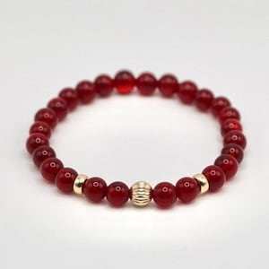 May include: A red gemstone bracelet with a gold bead accent. The bracelet is made of small, round beads and is strung on a stretchy cord.