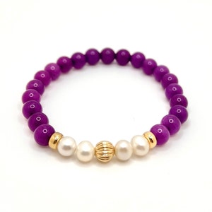 May include: A purple and white beaded bracelet with gold accents. The bracelet is made with round beads and has a gold bead in the center.