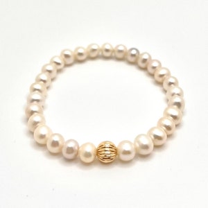Puede incluir: Un delicado brazalete con perlas blancas cremosas. Una sola cuenta dorada texturizada añade un toque de elegancia. El brazalete se muestra sobre un fondo blanco, resaltando su belleza simple y su versatilidad.