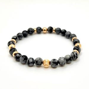 May include: A black and grey gemstone bracelet with gold accents. The bracelet is made of round beads and has a gold bead in the center.