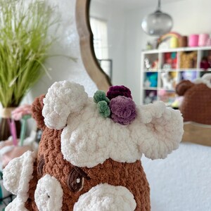 May include: A brown and white crocheted stuffed animal with a white fluffy face and a crown of purple, green and white flowers on its head.