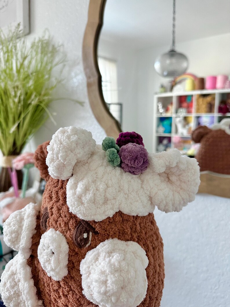 May include: A brown and white crocheted stuffed animal with a white fluffy face and a crown of purple, green and white flowers on its head.