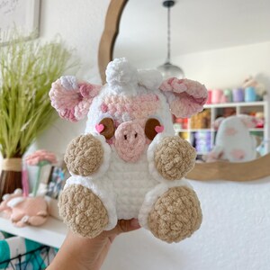May include: A crocheted stuffed animal pig with pink and white fur, brown eyes, and brown hooves. The pig is holding its hands in front of its face.