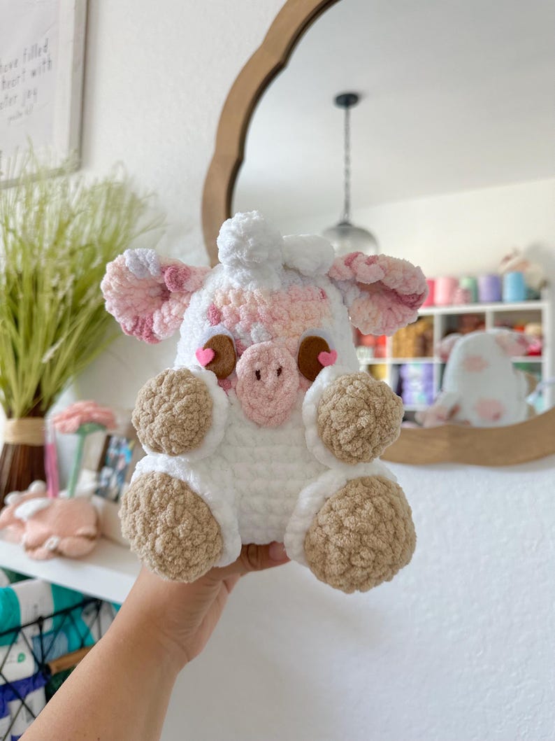 May include: A crocheted stuffed animal pig with pink and white fur, brown eyes, and brown hooves. The pig is holding its hands in front of its face.