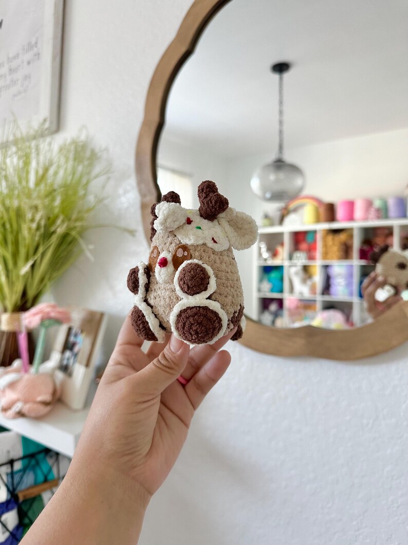May include: A crocheted reindeer ornament made with brown and white yarn. The reindeer has a brown nose and brown eyes. The ornament is decorated with white and red sprinkles.