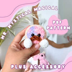 May include: A crocheted, circular accessory with a pink and lilac top, brown moustache, and white pom-pom paws. The text "MR. MYSTICAL MAGICAL PDF PATTERN PLUS ACCESSORY" is displayed in a pink font.