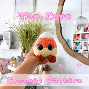 May include: A handmade crochet candy corn figure with large, glittery eyes and small brown feet. The candy corn is white, orange, and yellow. The text "Tom Corn" and "Crochet Pattern" are displayed in pink.