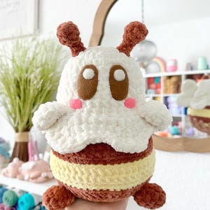 May include: A crocheted bee plush toy with brown and yellow stripes, white wings, and brown antennae. The bee has large brown eyes and pink cheeks.